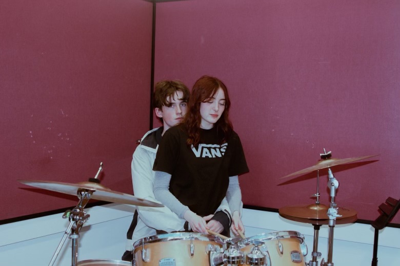 April and Lucien seated by a drum kit in a rehearsal room